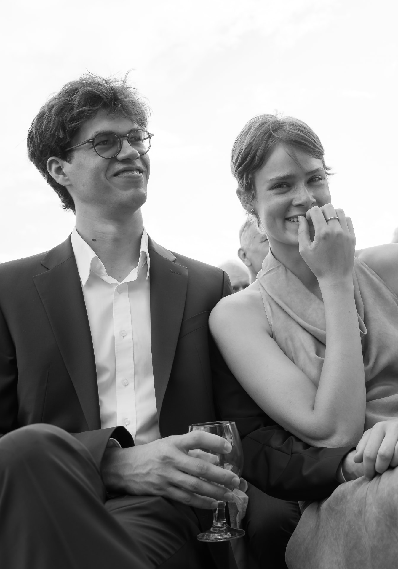 Black and white candid of a smiling couple seated at a wedding, wine glass in hand