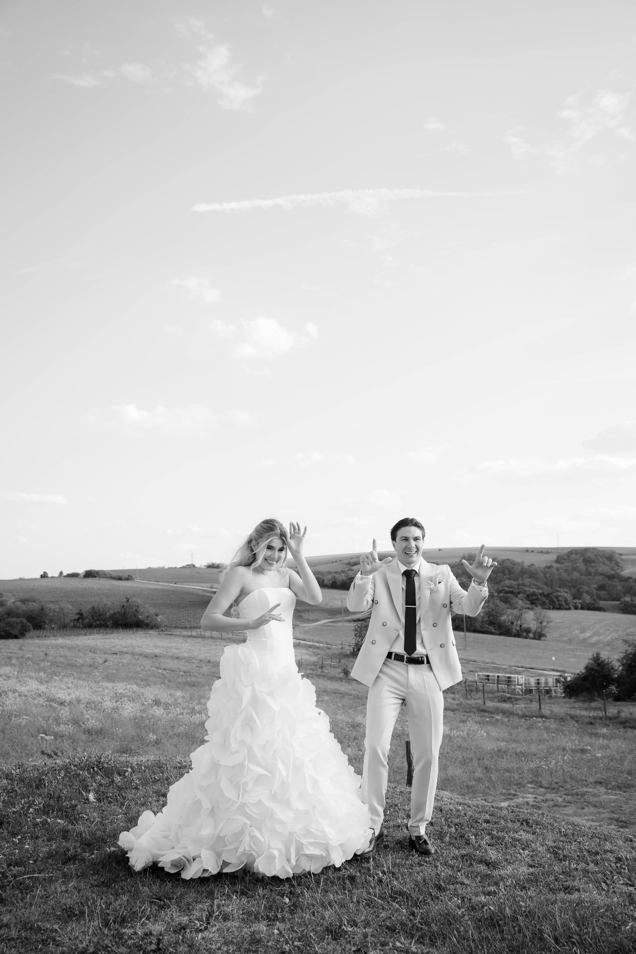 Bride and groom making playful hand gestures on a hillside meadow, black and white