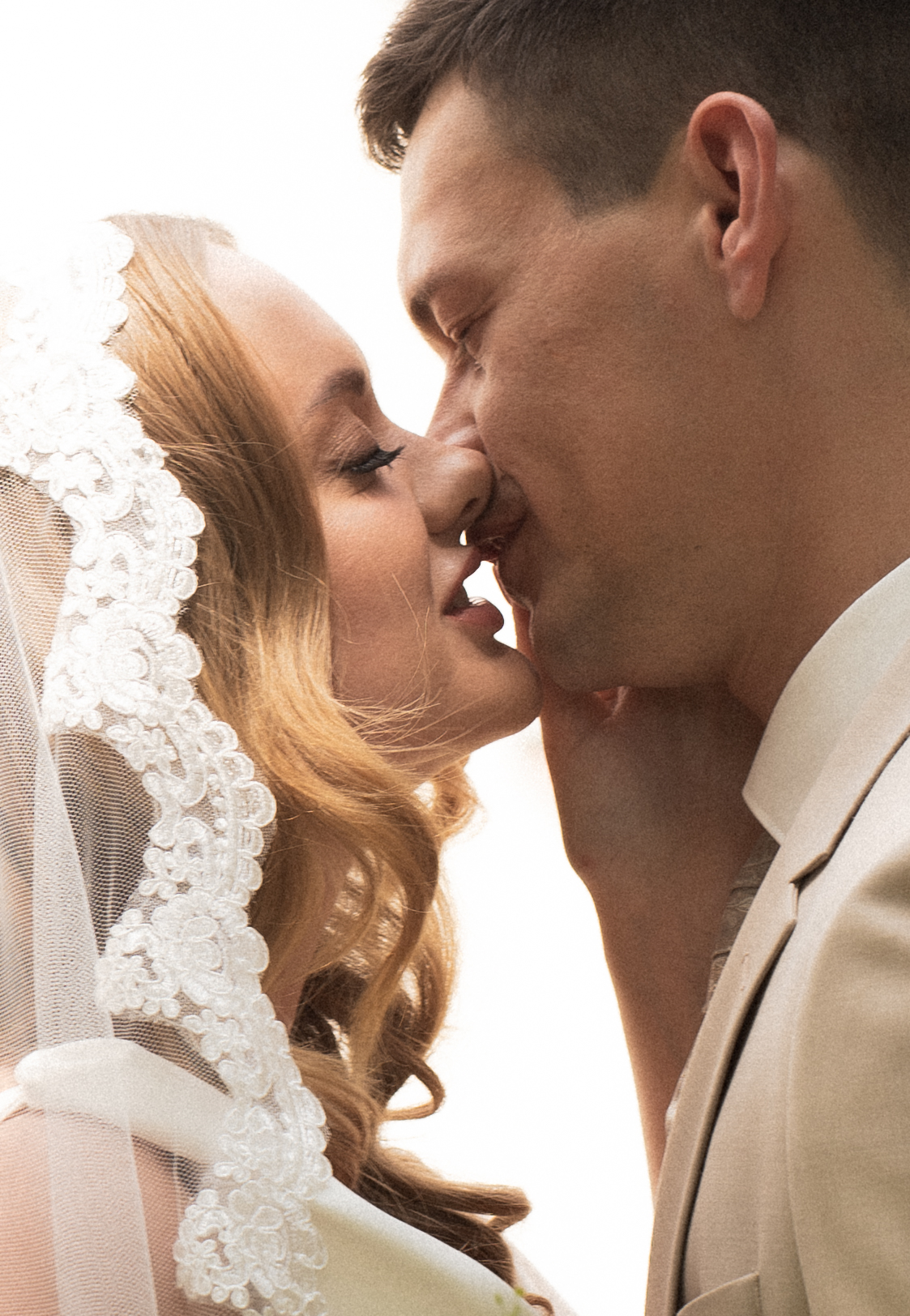 Backlit close-up of a bride in a lace veil kissing the groom