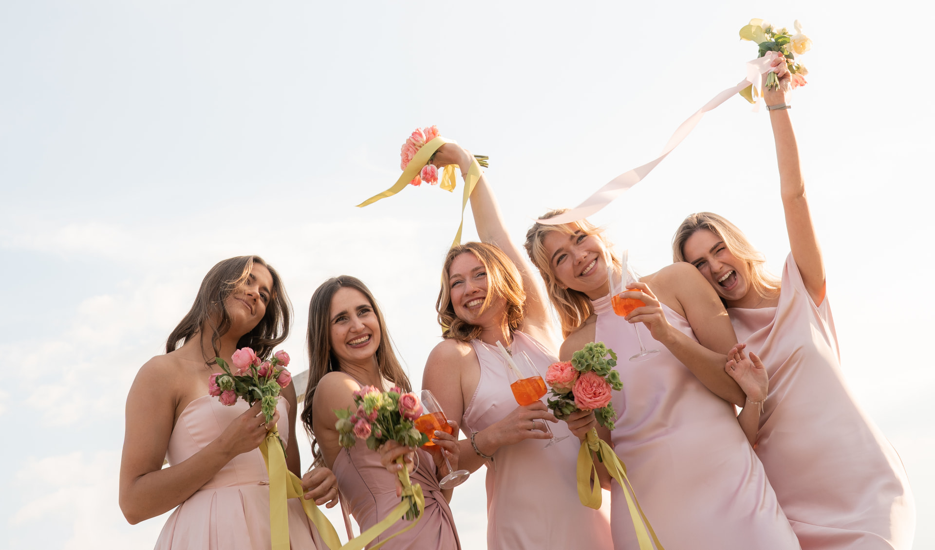 Bridesmaids in pink dresses celebrating with flowers and drinks against a bright sky