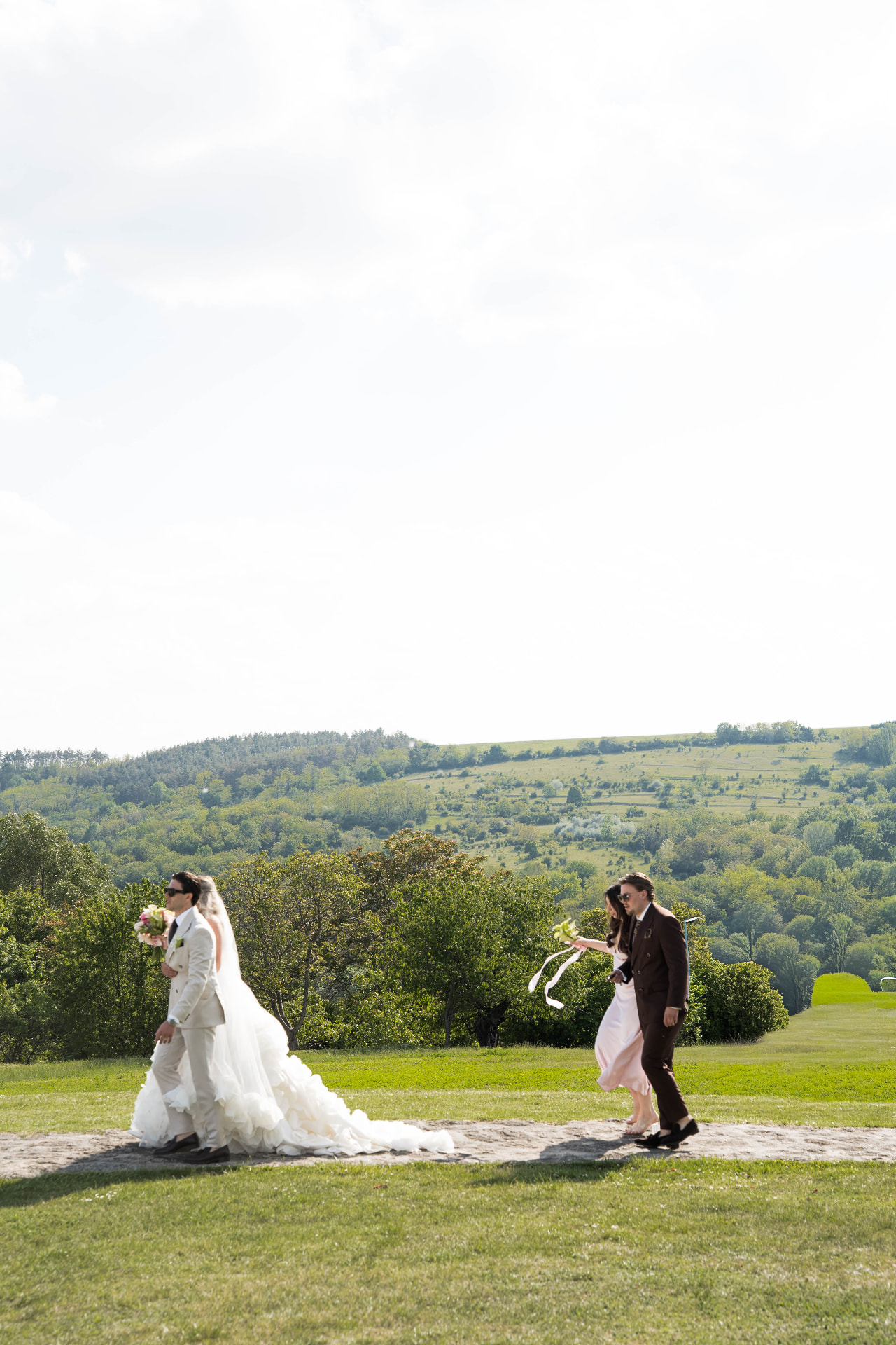 Bride and groom walking across a sunlit hillside meadow with guests following