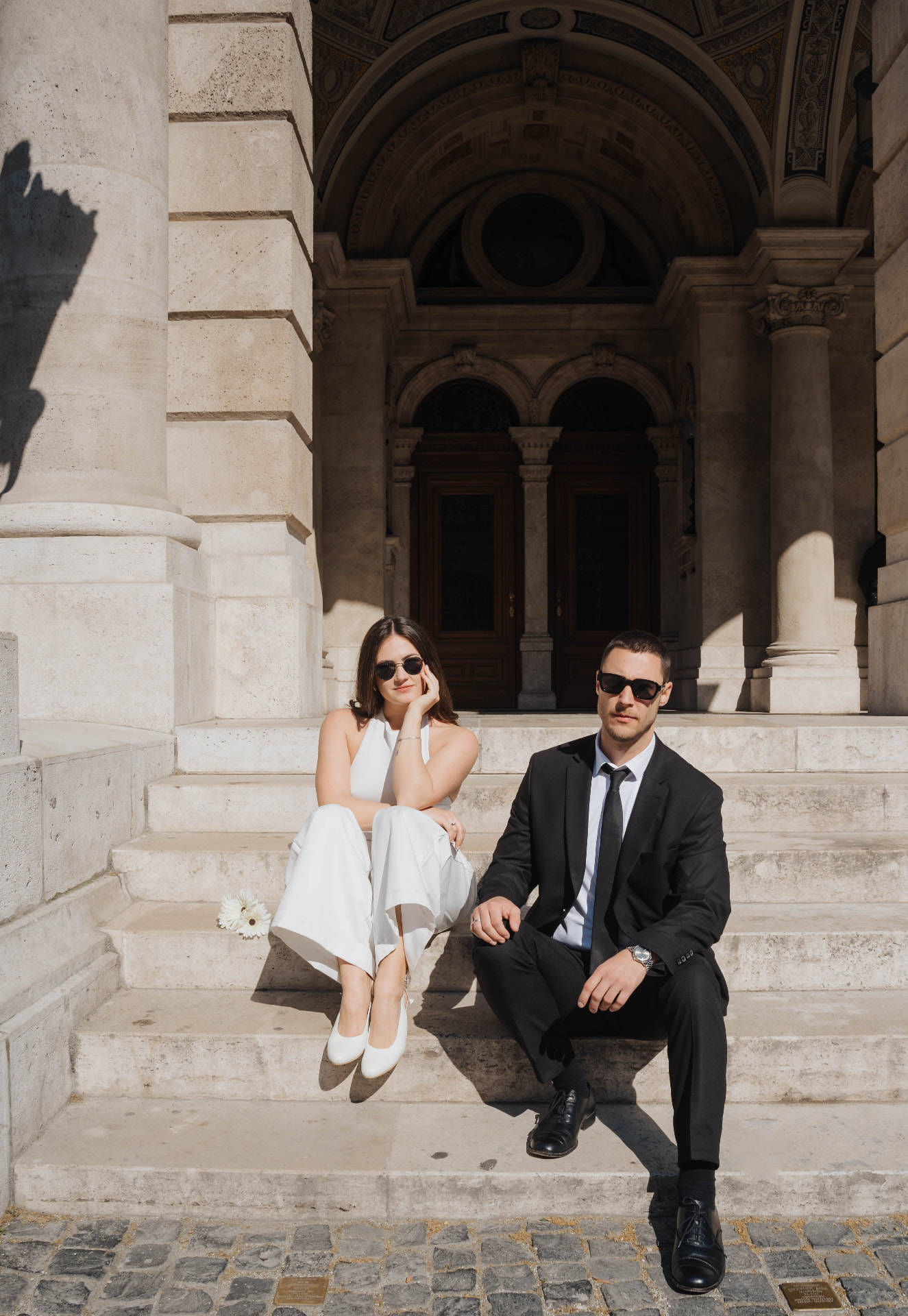 Bride and groom in sunglasses sitting on the steps of a grand building