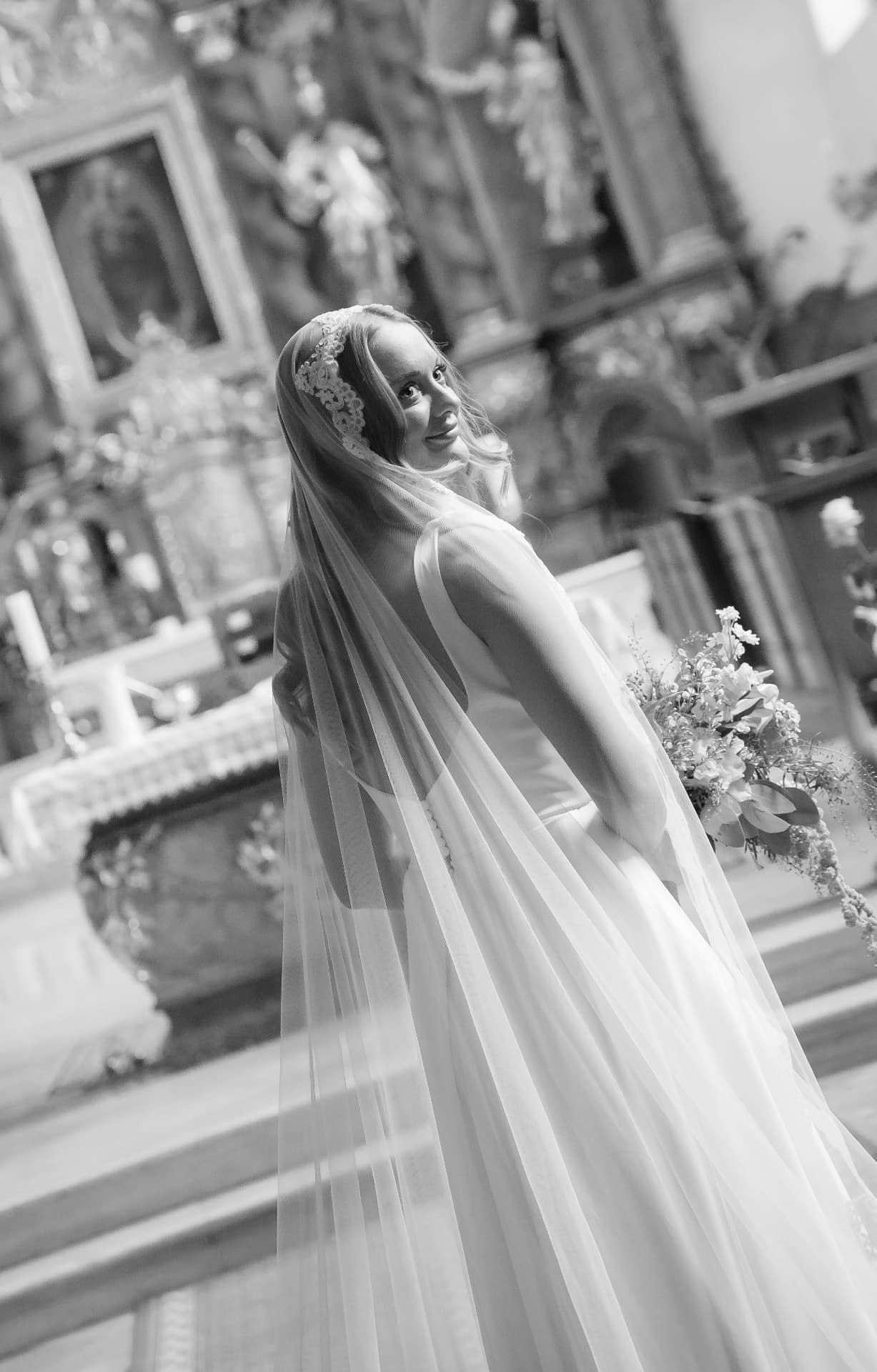 Bride in a long veil looking over her shoulder inside an ornate church, black and white