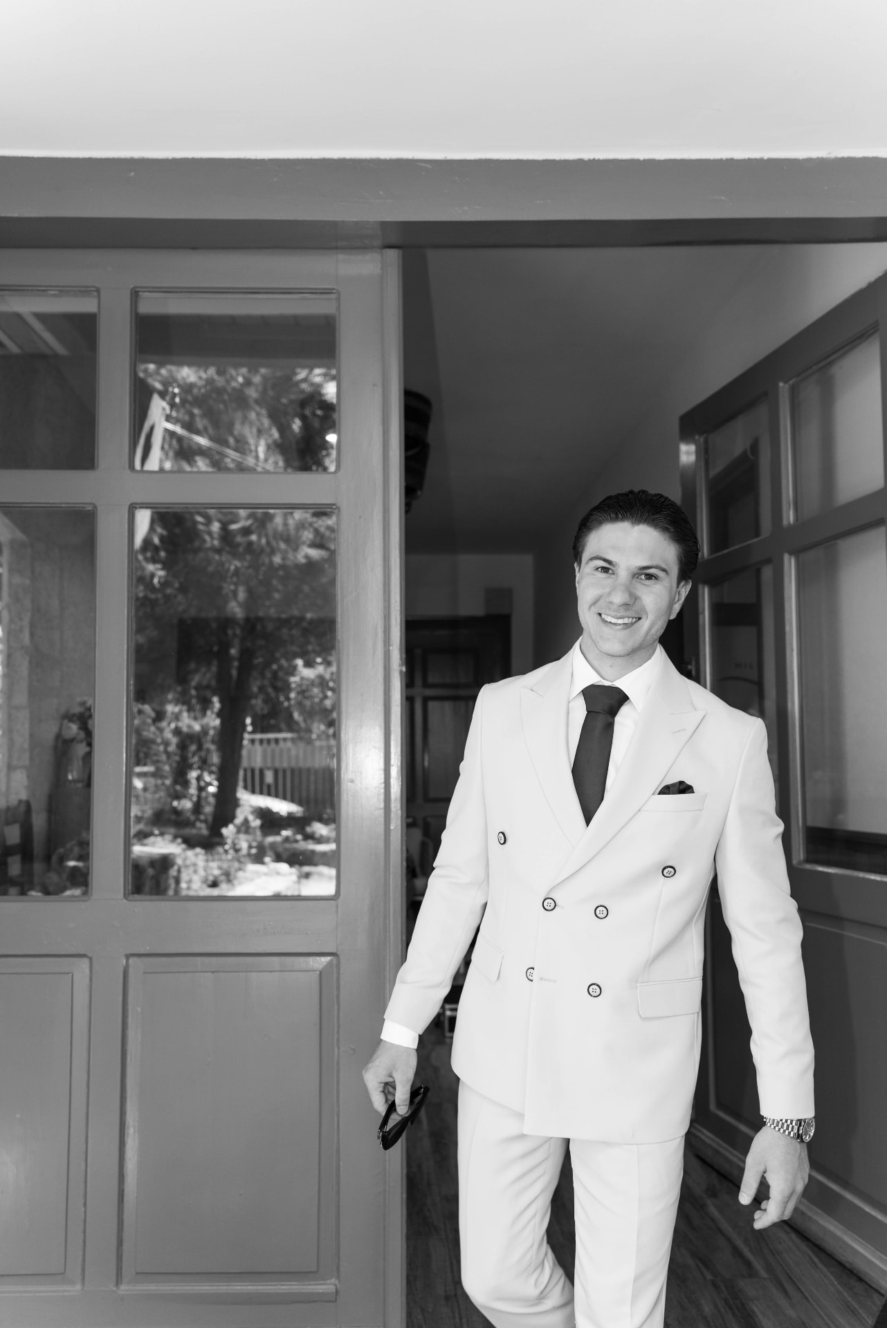 Groom in a white suit smiling in a doorway, black and white portrait